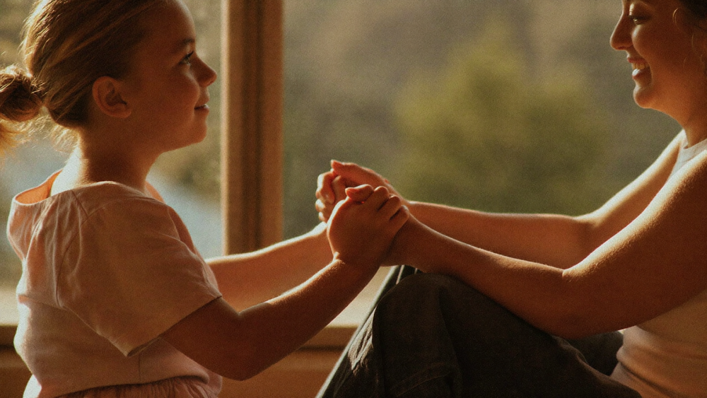 A daughter and her mother holding hands by a sunlit window